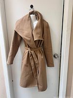 Full front view of the beige women's jacket hanging on a wall hook, showing the wide collar and belt.