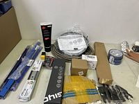 Overview of assorted automotive and tool-related items on table including clamps, cable coil, adhesive tube, tape, boxed parts, and cloth.