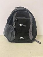 Front view of the Vale High Sierra backpack showing black and gray fabric with front zippered pocket, mesh side pocket, and logos.