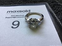 Front angle view of sterling silver ring with large clear faceted gemstone and detailed band on MaxSold paper.