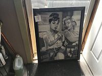 Front view of the large framed monochrome artwork showing tattooed Marilyn Monroe and Audrey Hepburn seated together, displayed near a door.
