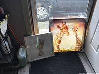 Two giraffe artworks shown near a window: a framed giraffe with floral crown and a canvas of two giraffes.