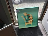 Square art print of Marilyn Monroe in a wicker chair with teal background, possibly with some light staining on the right side.