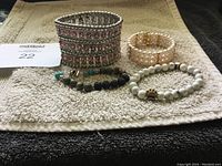 Four bracelets shown on a light colored cloth including a wide pink rhinestone cuff, white pearl stretch bracelet, multi-color beaded bracelet with turquoise and lava beads, and white stone bead bracelet with gold bead.