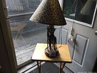Full view of the giraffe table lamp positioned on a small wooden table in front of a door and window. Shows the overall shape, wood-like giraffe figures, and leopard print lampshade.