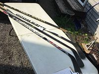 Four used right hand hockey sticks laid out on a white table outdoors. Sticks show wear, used black blades with visible scuffs.