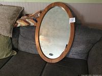 Oval oak framed hall mirror resting horizontally on couch cushions with reflective mirror glass showing some background.