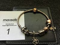 Photo showing the Pandora charm bracelet with its gold clasp and multiple charms laid on a black surface with a MaxSold catalog card.