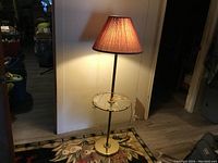 Pole lamp turned on showing lampshade and built-in glass table on metal pole and base.