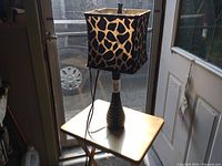 Full view of the table lamp with giraffe pattern shade illuminated and resting on a light wood square table against a door window.