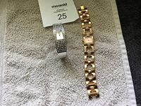 Two wristwatches placed on a towel, one silver-tone with black stones, the other gold-tone with rhinestones on the band.