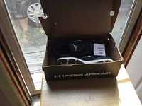 Open shoebox displaying the black Under Armour shoes with white soles inside the box, positioned near a window with natural light.