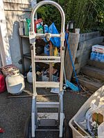 Front view of the Magliner aluminum hand truck showing the brand name, handlebar, and wheels. Background includes various garage and outdoor items.