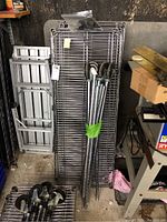 Photo showing 5 flat wire metal shelves stacked against a wall with 5 metal poles fitted with caster wheels bundled together and leaning against the shelves. Shelves appear rectangular with tubular framing.