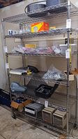 Full view of the stainless steel metal wire shelving unit with five shelves, stocked with various miscellaneous items. Showcases overall structure, shelving wire pattern, and wheels on bottom.