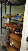 5-tier metal shelving unit holding various boxed model train sets and accessories; plastic bubble wrap and cardboard boxes stored on bottom shelf and floor around shelf.