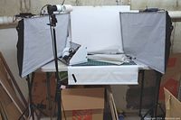 View of photography lighting setup including two softbox lights with bulbs on tripods, white backdrops, two umbrellas, cutting mats, and other accessories on a tabletop.