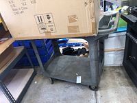 Side view of Rubbermaid rolling utility cart, showing two shelves and caster wheels, with a cardboard box placed on top.