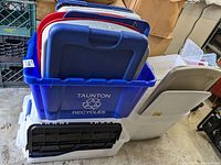 Stack of blue Taunton recycling bin and various colored lids including black, red, blue, gray, and white, plus white and translucent plastic bins with lids in garage setting.