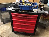 Front view of closed rolling tool chest showing black body with red drawers and organizing trays on top containing small hand tools and hardware.