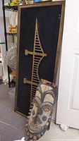 Both items together leaning against a door: the long rectangular framed woodcraft and the carved tribal mask in front.