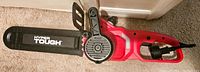 Side view of red and black Hyper Tough electric chainsaw showing the protective hand guard and the chain bar with brand name visible.