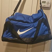 Front and side view of the large blue and black Nike duffle bag hanging by its shoulder strap, showing white Nike swoosh logo and black carrying handles.