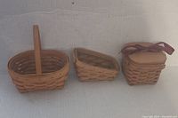 Three small Longaberger baskets: an oval handled basket, a small triangular basket, and a square basket with wooden lid and red ribbon.