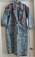 Full length front view of acid wash denim jacket with colorful geometric patterns on shoulders and sleeves, hanging on door.
