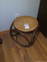 Photo of the stool showing top circular cane woven seat and rattan bent frame on wood floor near wall outlet.