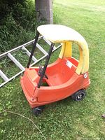 Side view of orange and yellow child's plastic car sitting on grass with a metal ladder in the background.