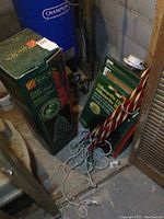 Fiber optic Christmas tree box, two lit candy cane decorations with lights, and four silhouette holiday lights stacked near a wall in basement setting.