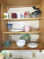 Full view inside cupboard showing various Corelle dishes, mugs, glass cups, bowls, and hand chopper