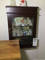 Front and angled view of dark brown wooden wall mount cabinet with glass door showing internal collectible glass figurines on two shelves.