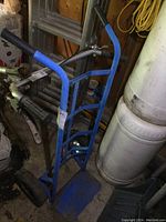 Blue metal utility dolly with solid wheels and black rubber handle grips. Visible signs of wear and paint chipping. Positioned near a ladder and other workshop items.