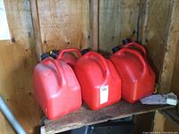 Five medium-sized red plastic fuel cans stacked in two rows on a wooden shelf inside a wood-paneled room. Cans have carry handles and black caps with yellow safety features.