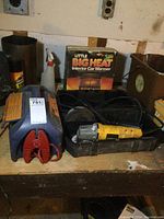 Booster pack (red and blue), boxed Big Heat car warmer, and Dewalt angle grinder in black case on wooden surface in workshop