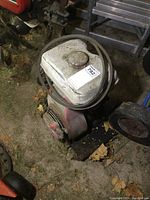Photo of the overall Honda motor showing white fuel tank and red motor base on dirt ground with some leaves.