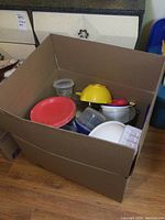 Photo of large cardboard box with assorted kitchen ware including plastic bowls, containers with lids, red plates, yellow colander, and a glass pitcher with green lid visible inside.