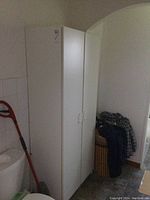 Front and side view of tall white pantry cabinet near a mop and basket.