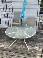 Round patio table with glass top and metal frame legs on wooden deck, two patio chairs and umbrella visible in background