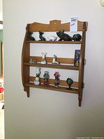 Front view of wooden wall rack mounted on wall with various small collectibles arranged on shelves including glass animal figurines, ceramic pitchers, miniature oil lamps, a small globe, and a framed picture.