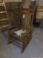 Solid wood larger size rocking chair with spindle details, dusty, with white and green leaf patterned cushion on seat, photographed in basement room.