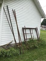 Metal scaffolding parts stacked against a white siding wall and grass, showing three main pieces: two frame assemblies and two narrow side supports on the left.