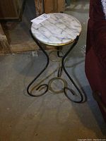 Side view of the round white marble top with grey veining on a black metal frame base with scroll design, showing the table in its entirety and signs of wear on marble and metal.
