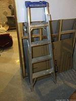 5 ft aluminum step ladder standing open and leaning against a cardboard background. Clear view of four wide aluminum steps and blue top cap.