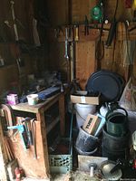 Overview of a corner of a wooden shed showing a wooden workbench with various small tools and containers on top. Below and around it are plastic milk crates, black bucket, and other supplies organized on shelves.