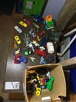 Overview photo showing arrangement of various toy cars, trucks, plastic figures and more scattered and in a box on the floor