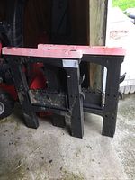 Two plastic resin folding sawhorses, black and red, leaning against a wall. Seen with dirt and paint wear.