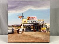 Full view of square canvas print showing cowboy trading post under cloudy sky with vintage cars and multiple signs including Coca-Cola and Conoco